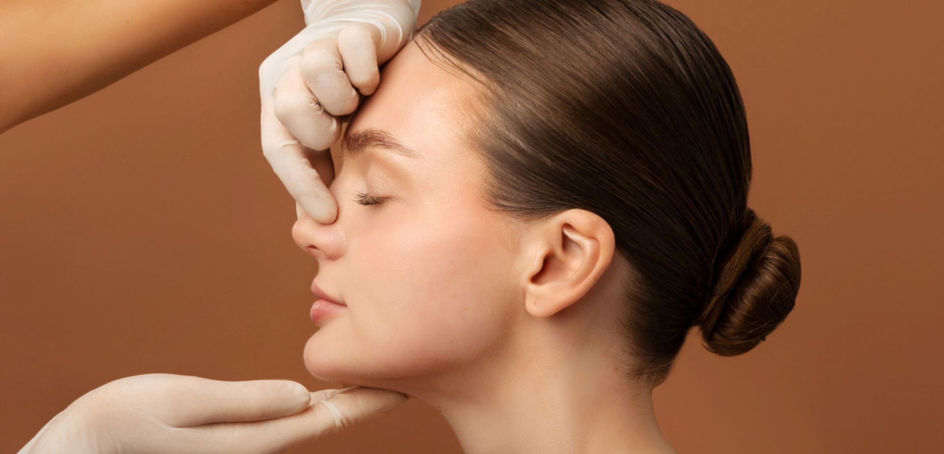 gloved-hands-assessing-a-woman's-nose-against-a-brown-background